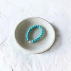 A turquoise beaded bracelet with a small silver charm, displayed inside a small speckled ceramic dish on a textured white surface.