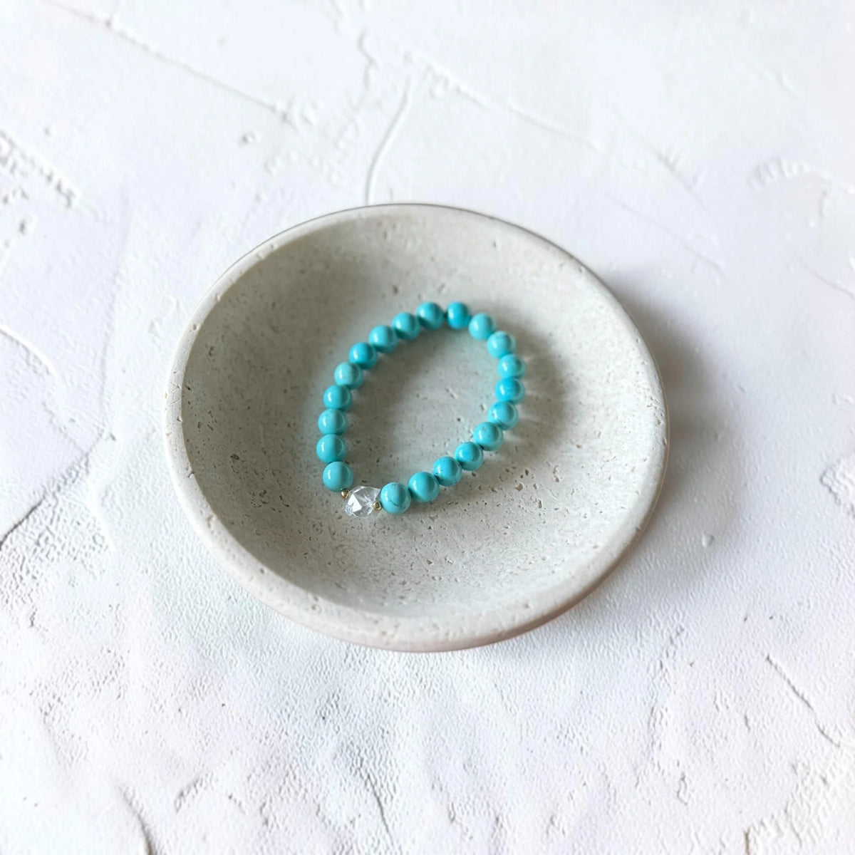 A turquoise beaded bracelet with a small silver charm, displayed inside a small speckled ceramic dish on a textured white surface.