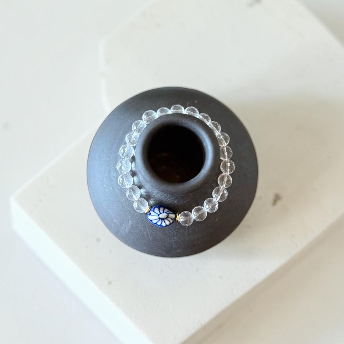 A beaded bracelet with clear faceted beads and a small blue-and-white floral accent bead, draped around a matte black ceramic vase on a white surface.