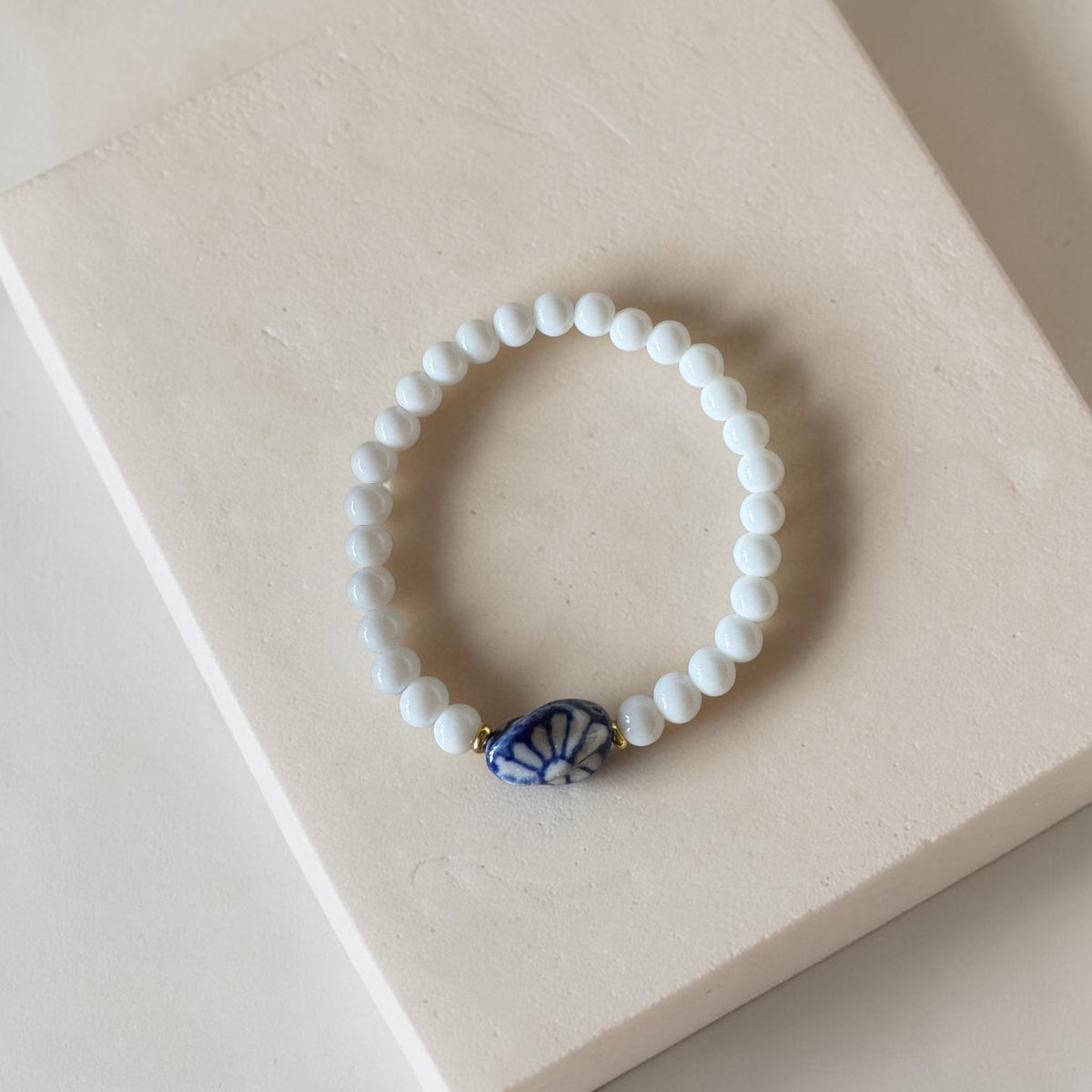 A beaded stretch bracelet with white round beads and a central blue and white patterned shell-shaped bead, displayed on a light beige textured surface.