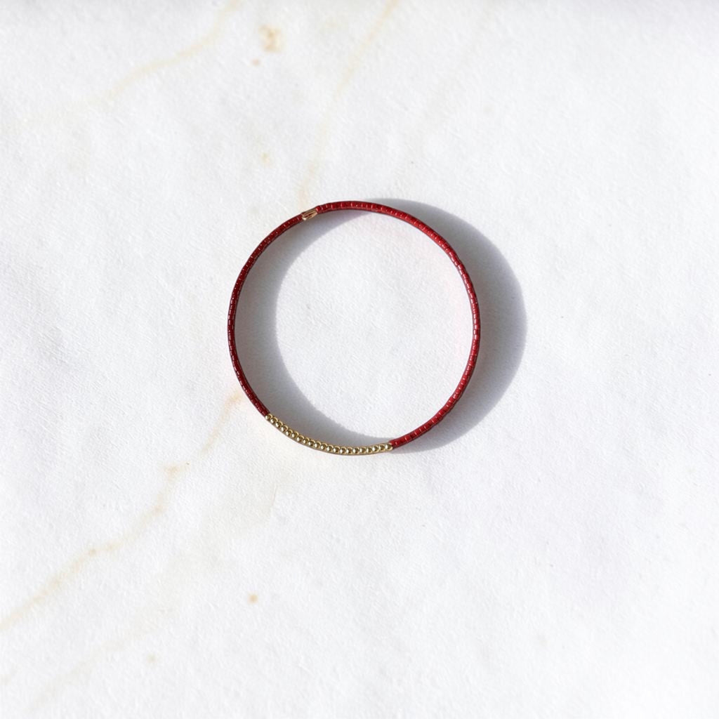 A thin red leather bangle bracelet with a small gold bead accent on one side, displayed on a white marble surface.
