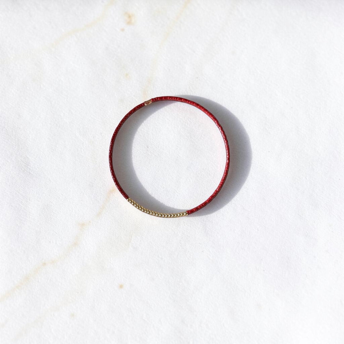 A thin red leather bangle bracelet with a small gold bead accent on one side, displayed on a white marble surface.