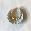 A beaded bracelet with cream, white, and pale swirl beads and a small gold accent bead, displayed on a small scalloped ceramic dish over a white fabric background.