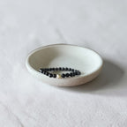 A beaded stretch bracelet with black round beads and a small gold accent bead, displayed inside a small white ceramic dish on a textured white fabric background.