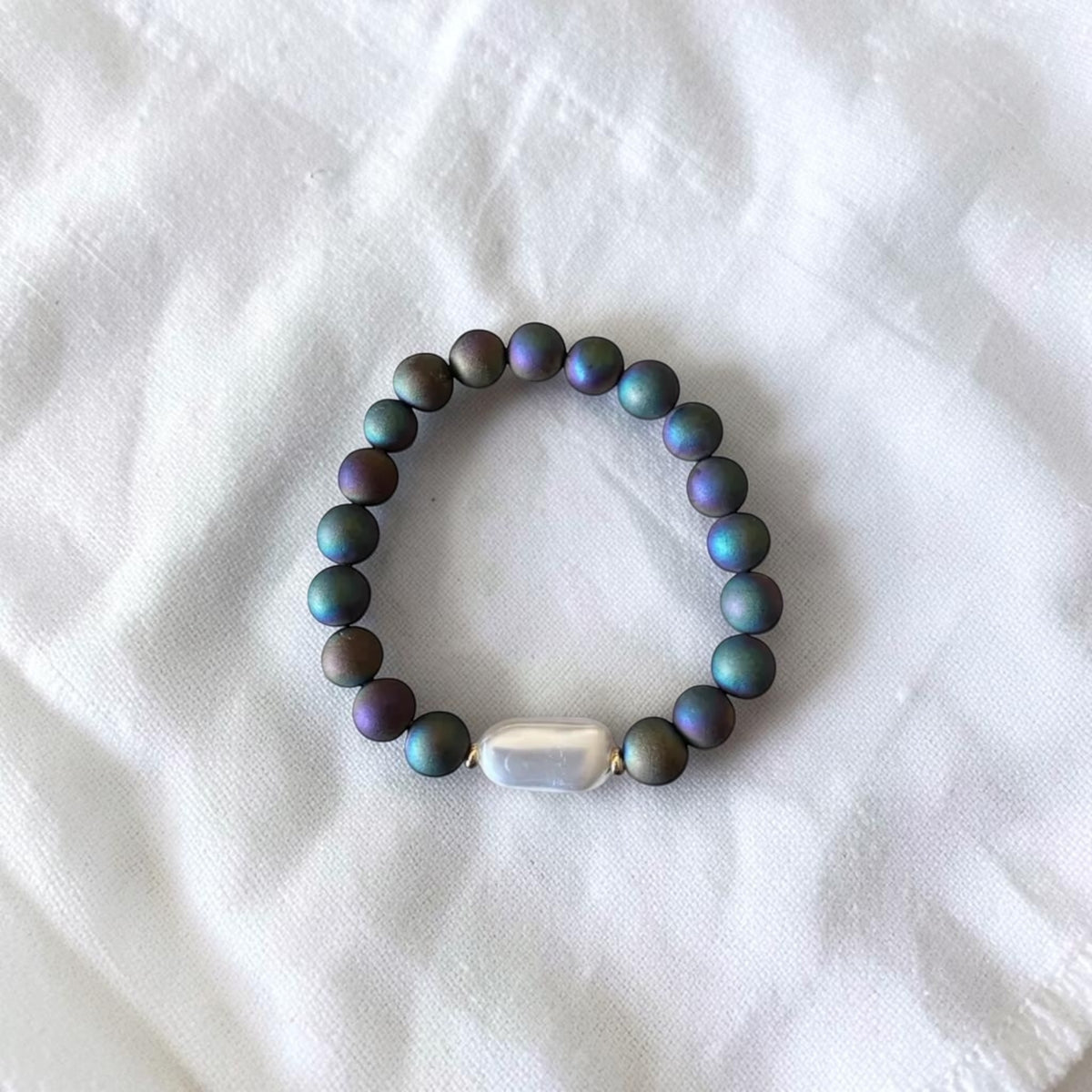 A beaded stretch bracelet featuring iridescent dark gray beads with blue and purple shifts, accented by a central white baroque pearl and gold spacer, displayed on a white fabric background.