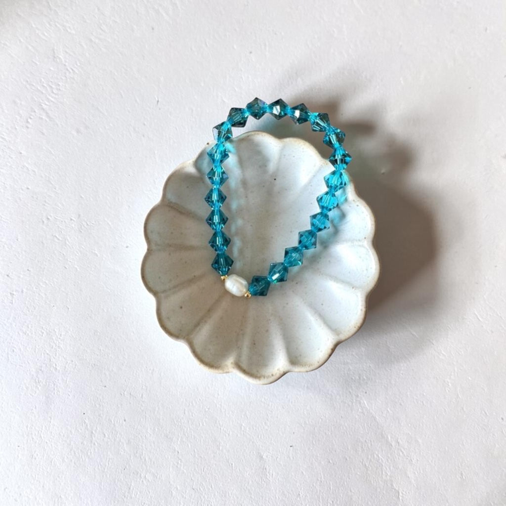A beaded stretch bracelet made of faceted turquoise-blue beads with a small white pearl accent, displayed on a white scalloped dish against a textured white background.