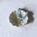 A light blue beaded bracelet with a small gold bow charm, displayed on a small white ceramic dish against a textured white background.