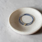 A pair of navy blue wire bracelets with small gold bead accents, displayed inside a round, textured off-white ceramic dish on a neutral gray surface.