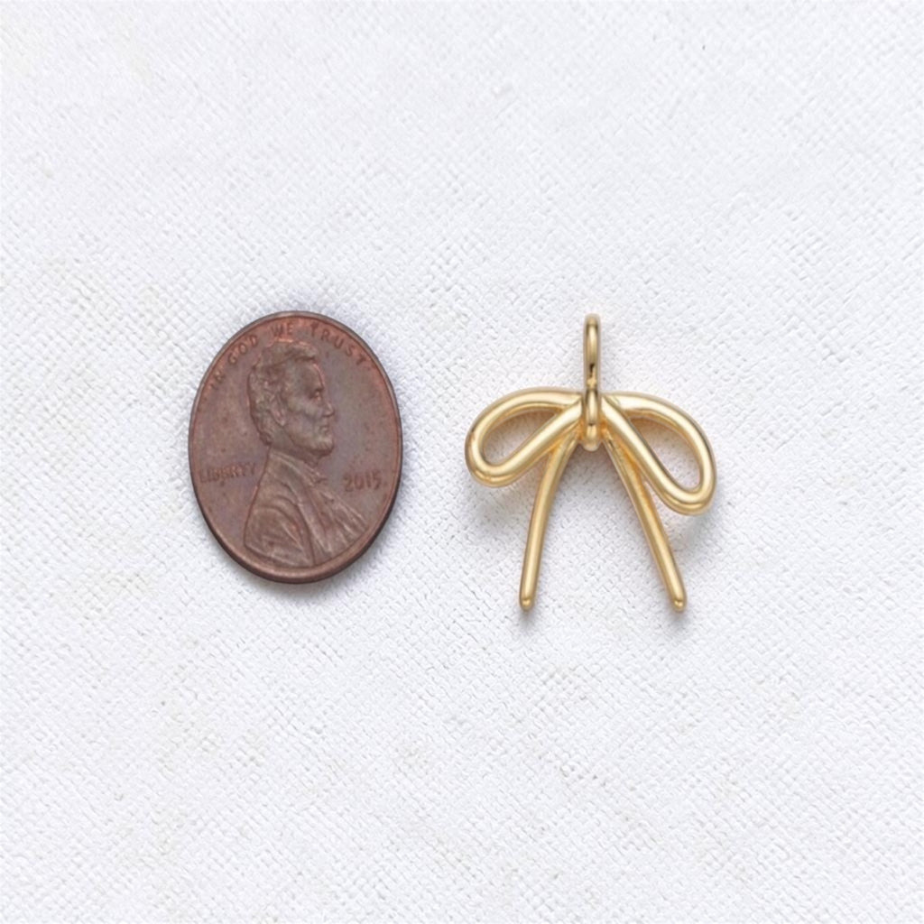 A small gold bow-shaped jewelry charm or pendant shown next to a U.S. penny for scale on a white textured background.