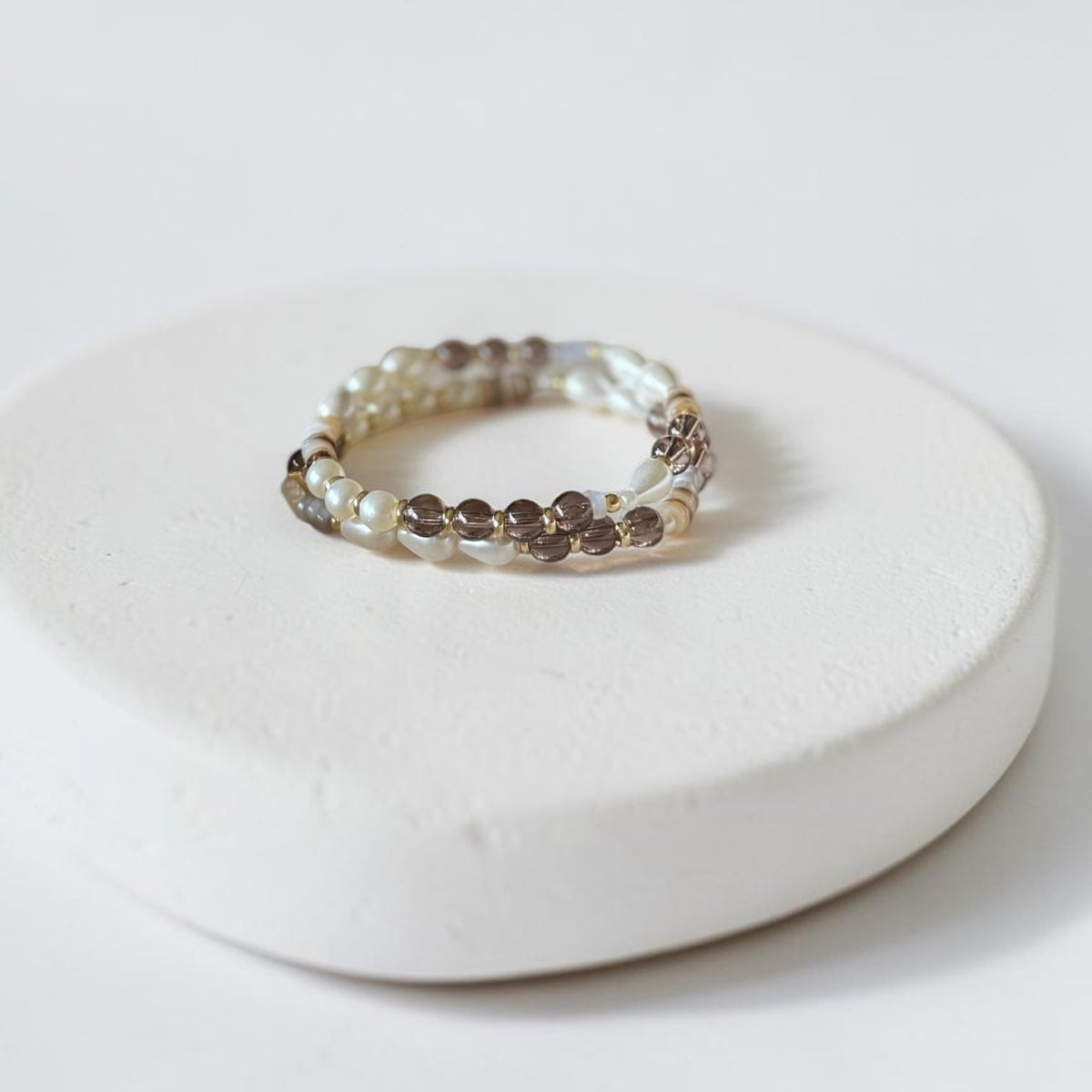 A beaded stretch bracelet with small brown and cream beads, displayed on a round white ceramic riser against a clean white background.