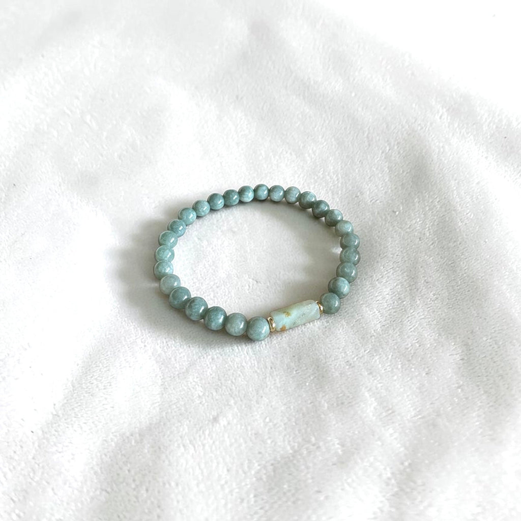 A beaded stretch bracelet with light blue-green round beads and a small white stone accent, resting on a white textured fabric background.
