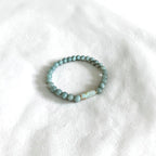 A beaded stretch bracelet with light blue-green round beads and a small white stone accent, resting on a white textured fabric background.