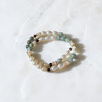 A pair of beaded stretch bracelets with white pearls, light blue beads, and small brown beads, displayed on a white marble surface.