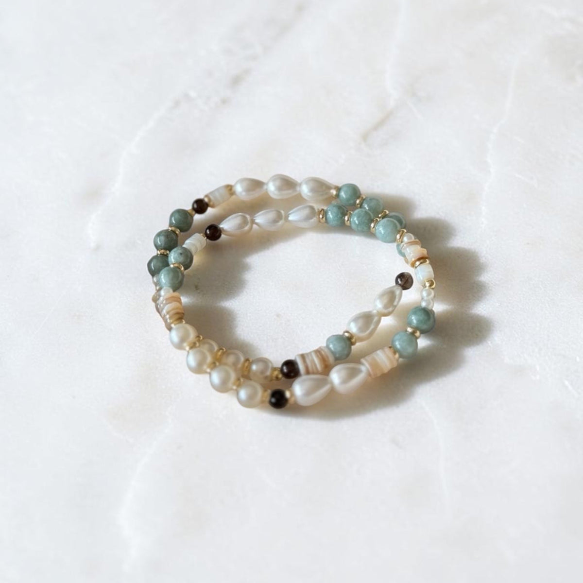 A pair of beaded stretch bracelets with white pearls, light blue beads, and small brown beads, displayed on a white marble surface.