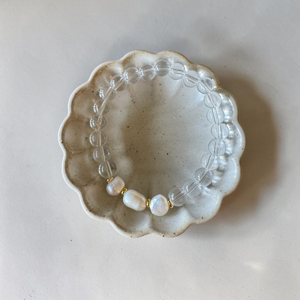 A beaded stretch bracelet with clear round beads and three white baroque pearls separated by small gold spacers, displayed on a scalloped ceramic dish against a light beige background.