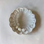 A beaded stretch bracelet with clear round beads and three white baroque pearls separated by small gold spacers, displayed on a scalloped ceramic dish against a light beige background.