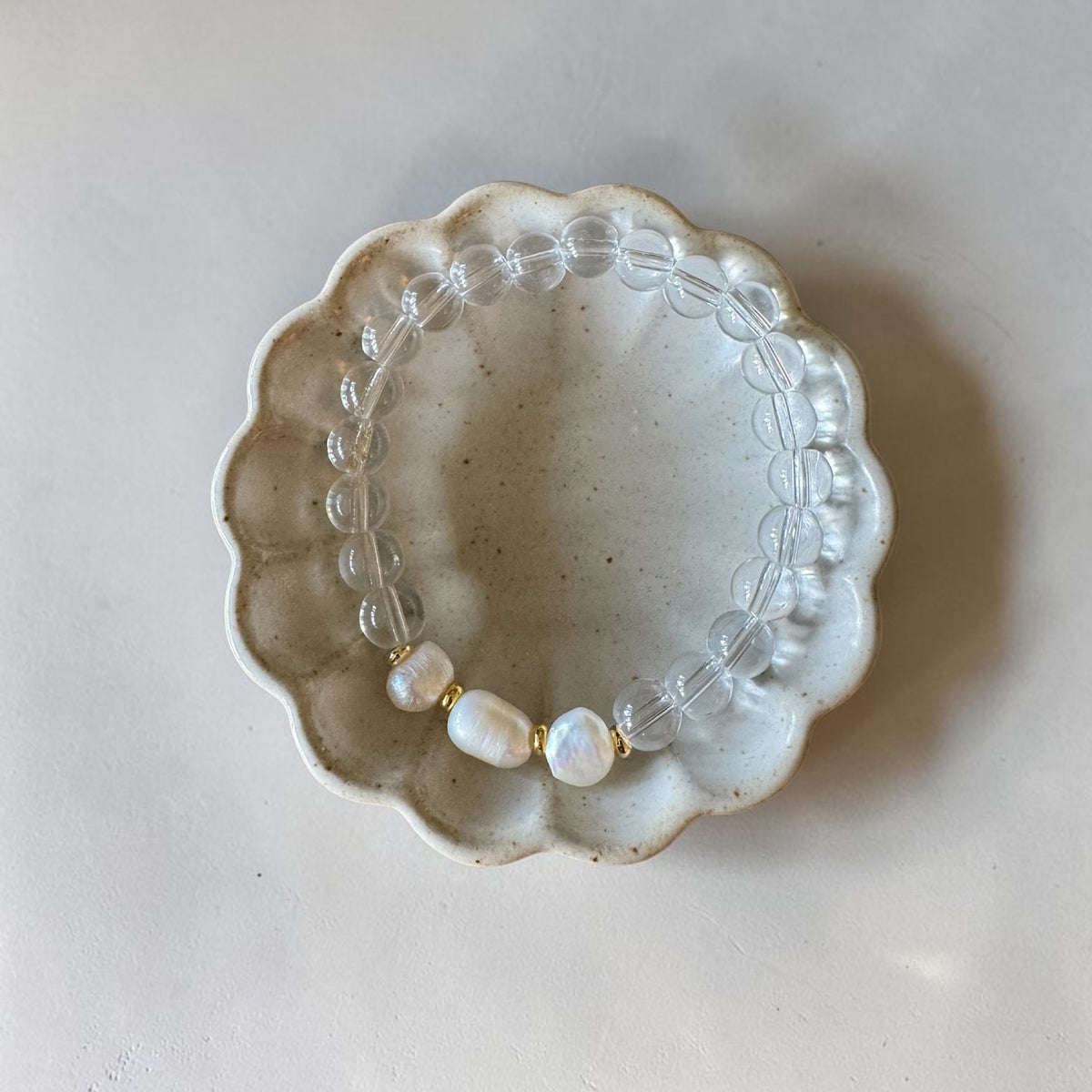 A beaded stretch bracelet with clear round beads and three white baroque pearls separated by small gold spacers, displayed on a scalloped ceramic dish against a light beige background.