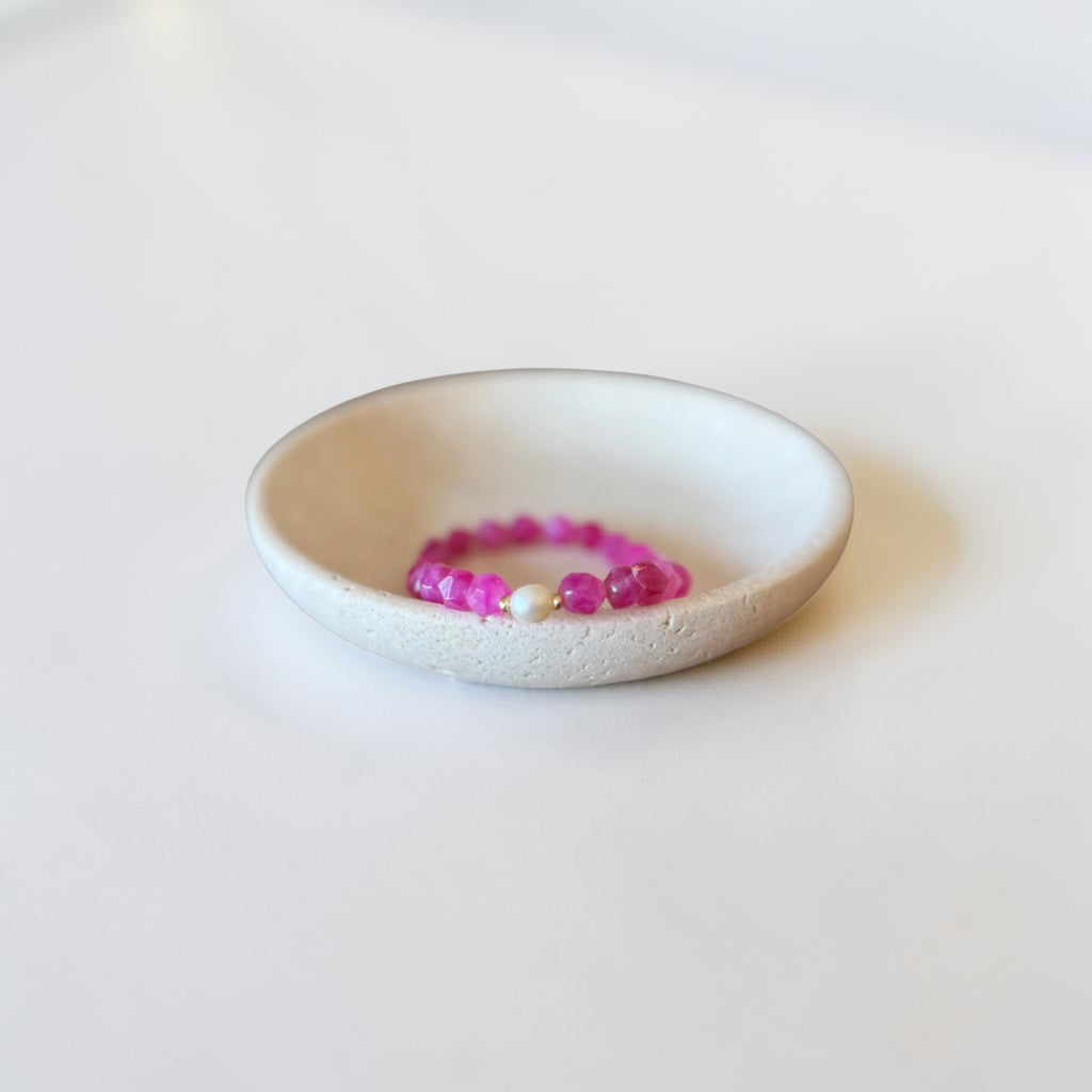 A small round ceramic dish holding a beaded stretch bracelet made of bright pink and purple beads with a single white bead accent.