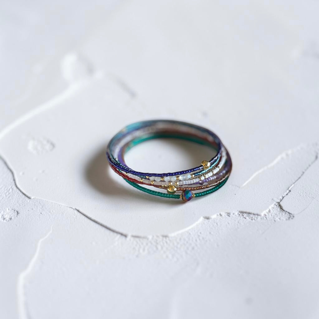 A delicate multi-layered ring with colorful glass strands, tiny white pearls, and small gold beads, resting on a textured white surface.