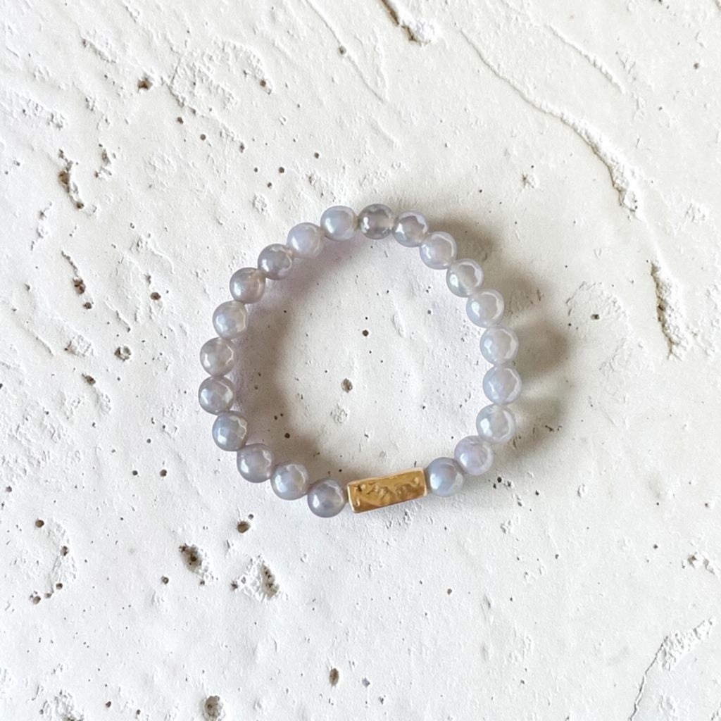A beaded stretch bracelet made of translucent light gray beads with a small gold rectangular accent, photographed on a textured off-white surface.