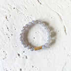 A beaded stretch bracelet made of translucent light gray beads with a small gold rectangular accent, photographed on a textured off-white surface.