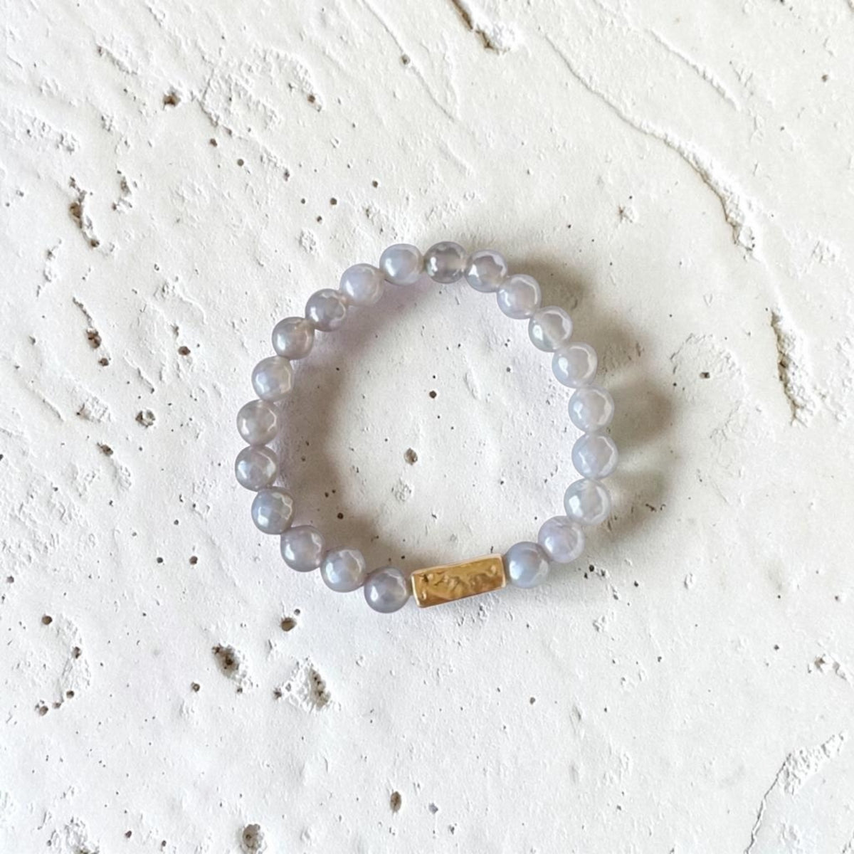 A beaded stretch bracelet made of translucent light gray beads with a small gold rectangular accent, photographed on a textured off-white surface.
