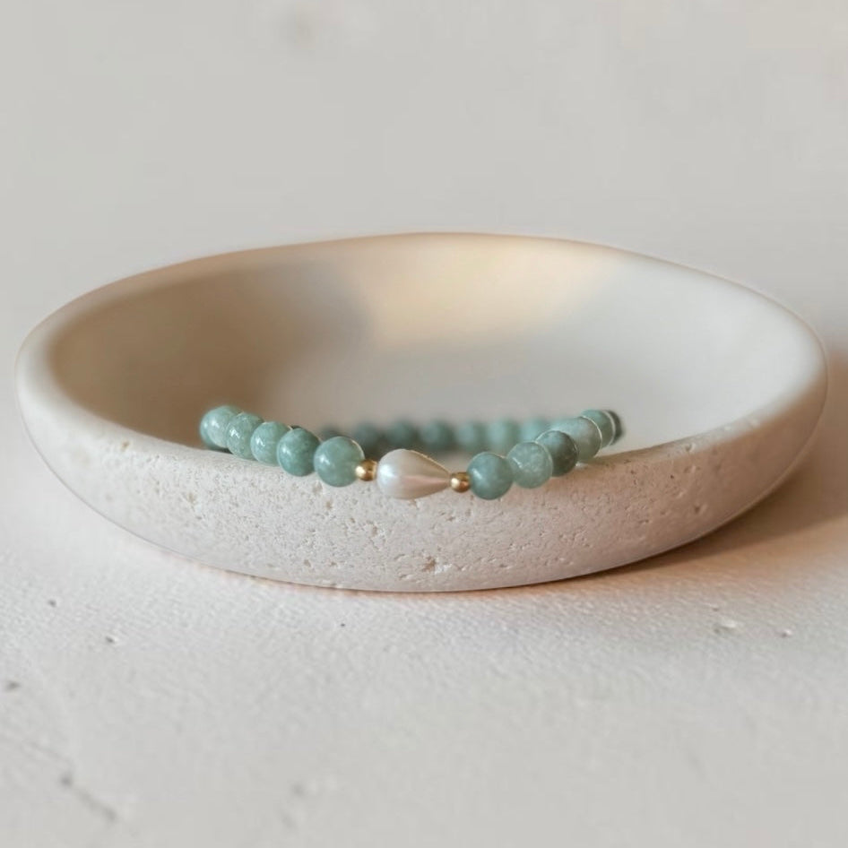 A beaded bracelet with  green round beads and a central white pearl-like accent, displayed in a shallow off-white ceramic dish on a textured light surface.