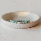A beaded bracelet with  green round beads and a central white pearl-like accent, displayed in a shallow off-white ceramic dish on a textured light surface.