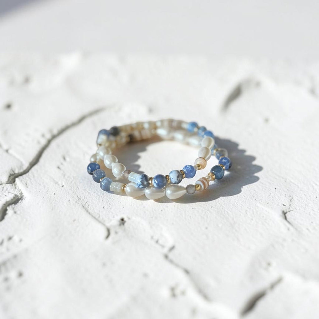 Two bracelets stacked on a white texture background. Blue Fire Agate Beads with a mixture of other accents. Pearls, Gold Plated Spacers.