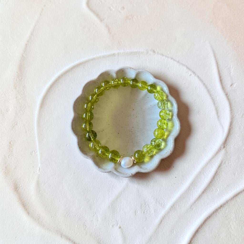 A beaded stretch bracelet made of translucent light green gemstone beads with a small white pearl accent, displayed on a small white scalloped dish over a textured beige surface.