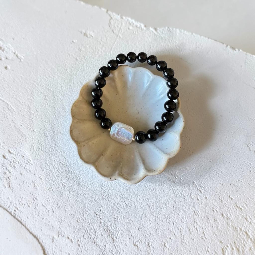 A beaded stretch bracelet with glossy black round beads and a central iridescent white bead, displayed on a scalloped cream ceramic dish against a textured off-white background.