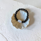 A beaded stretch bracelet with glossy black round beads and a central iridescent white bead, displayed on a scalloped cream ceramic dish against a textured off-white background.
