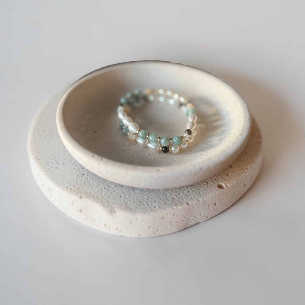 A beaded stretch bracelet with light green jade and white pearls sits in the shallow center of a small ceramic dish, which is placed on top of a larger round textured ceramic tray against a neutral light gray background.
