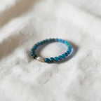 A beaded stretch bracelet with blue and teal beads, a small white bead, and a tiny gold spacer, resting on a textured off-white linen fabric.