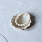 A beaded bracelet made of round, pale cream-colored beads, displayed inside a small, scalloped ceramic dish on a textured light gray surface.