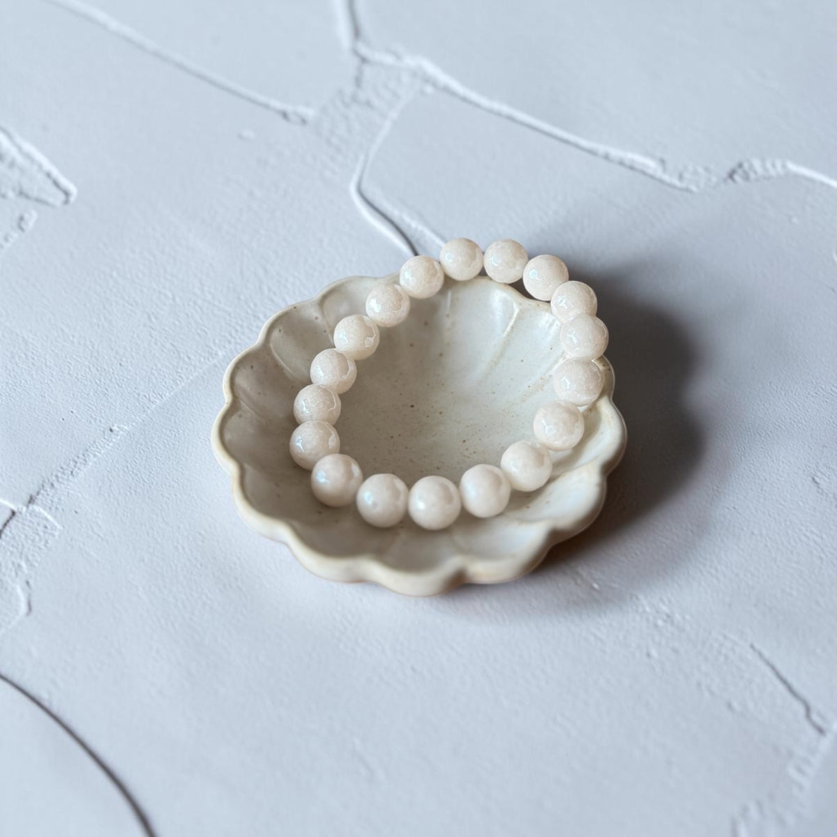 A beaded bracelet made of round, pale cream-colored beads, displayed inside a small, scalloped ceramic dish on a textured light gray surface.
