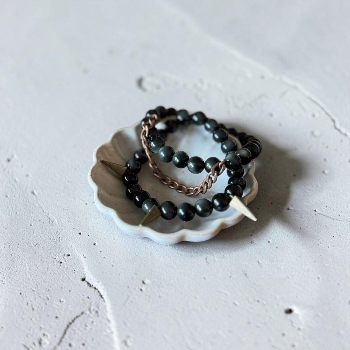 Two beaded bracelets displayed on a small white scalloped dish. The bracelets feature matte black beads, a dark metallic bronze chain, and a white tooth accent.