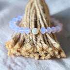 A beaded bracelet with translucent pale lavender beads and a central iridescent white bead, displayed on a textured beige jute stand against a soft white fabric background.