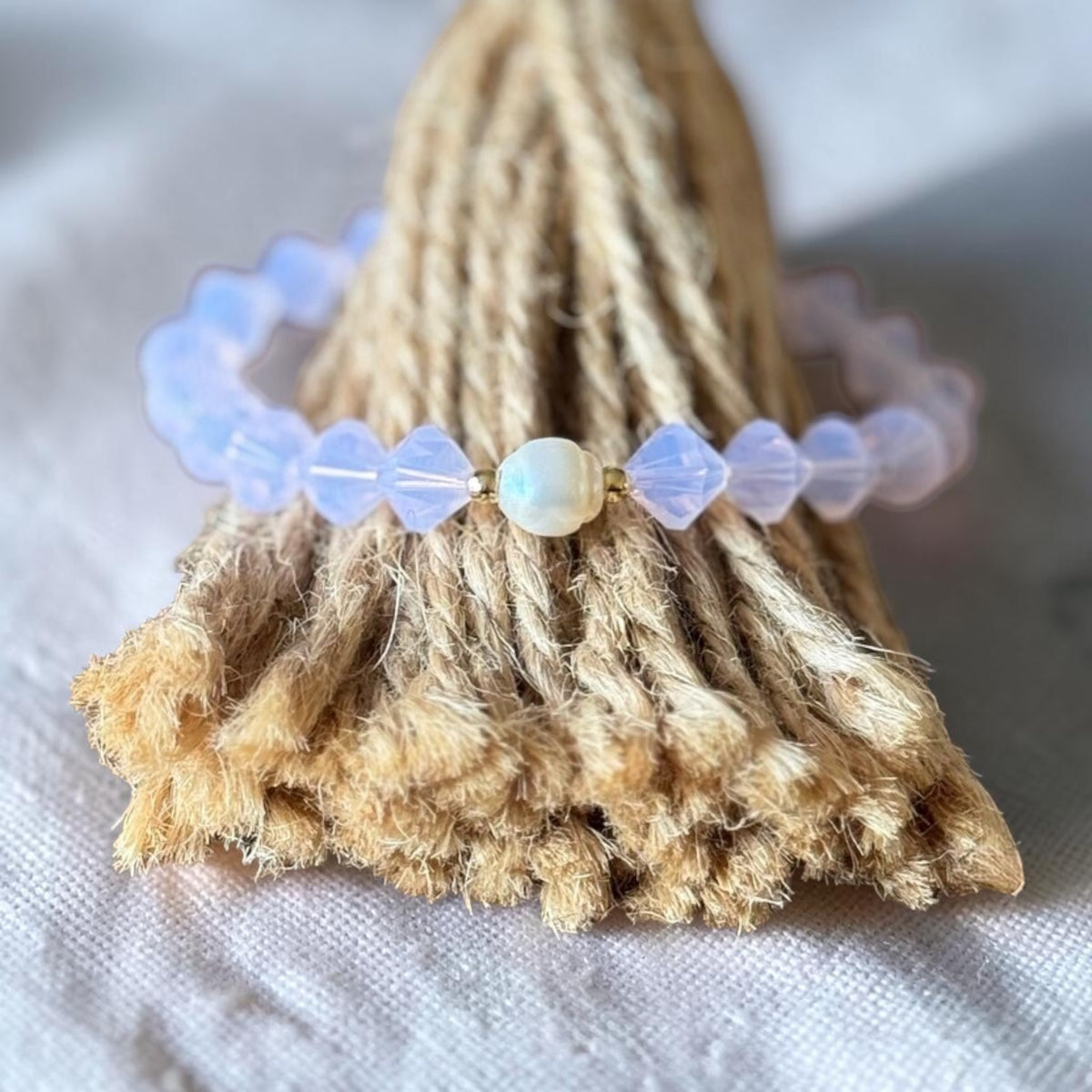 A beaded bracelet with translucent pale lavender beads and a central iridescent white bead, displayed on a textured beige jute stand against a soft white fabric background.