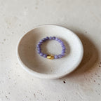 A beaded stretch ring with light purple lavender beads and a small gold metal accent, displayed inside a small speckled ceramic dish on a textured beige surface.