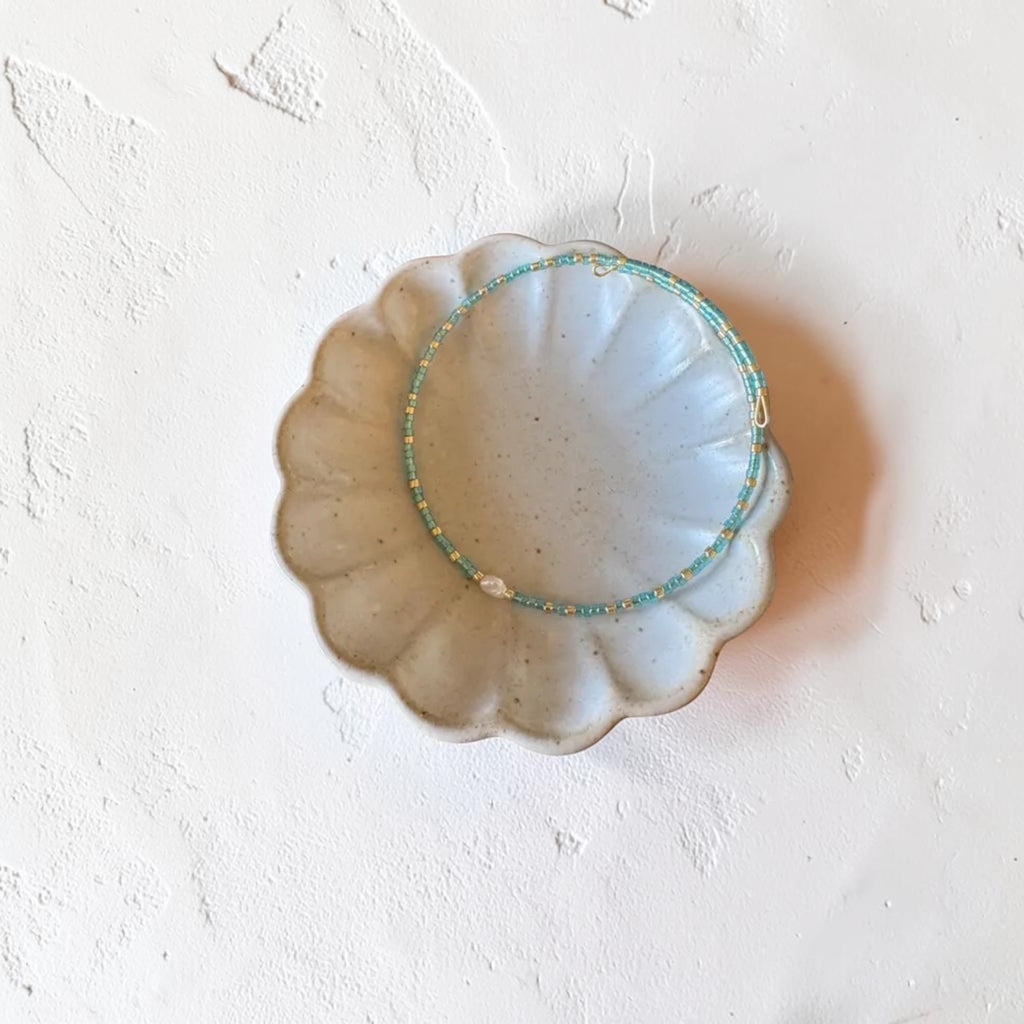 A delicate beaded bracelet with turquoise blue, gold, and white beads, displayed on a scalloped ceramic dish against a textured white background.