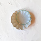 A delicate beaded bracelet with turquoise blue, gold, and white beads, displayed on a scalloped ceramic dish against a textured white background.