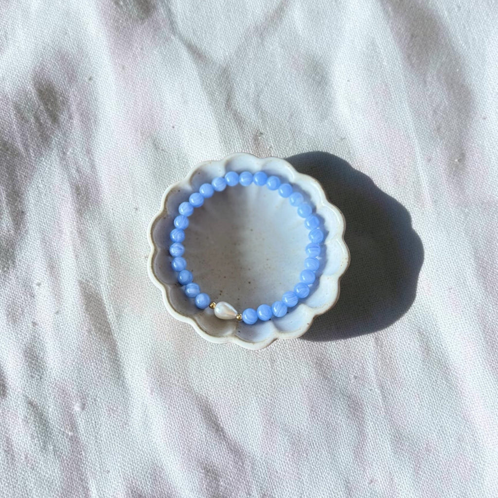 A light blue beaded bracelet displayed inside a small white scalloped dish on a textured white fabric background.