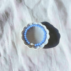 A light blue beaded bracelet displayed inside a small white scalloped dish on a textured white fabric background.