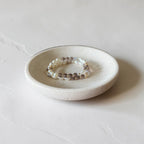 A beaded stretch bracelet with white pearls and brown gemstone beads resting on a small, round, textured ceramic dish against a light gray background.