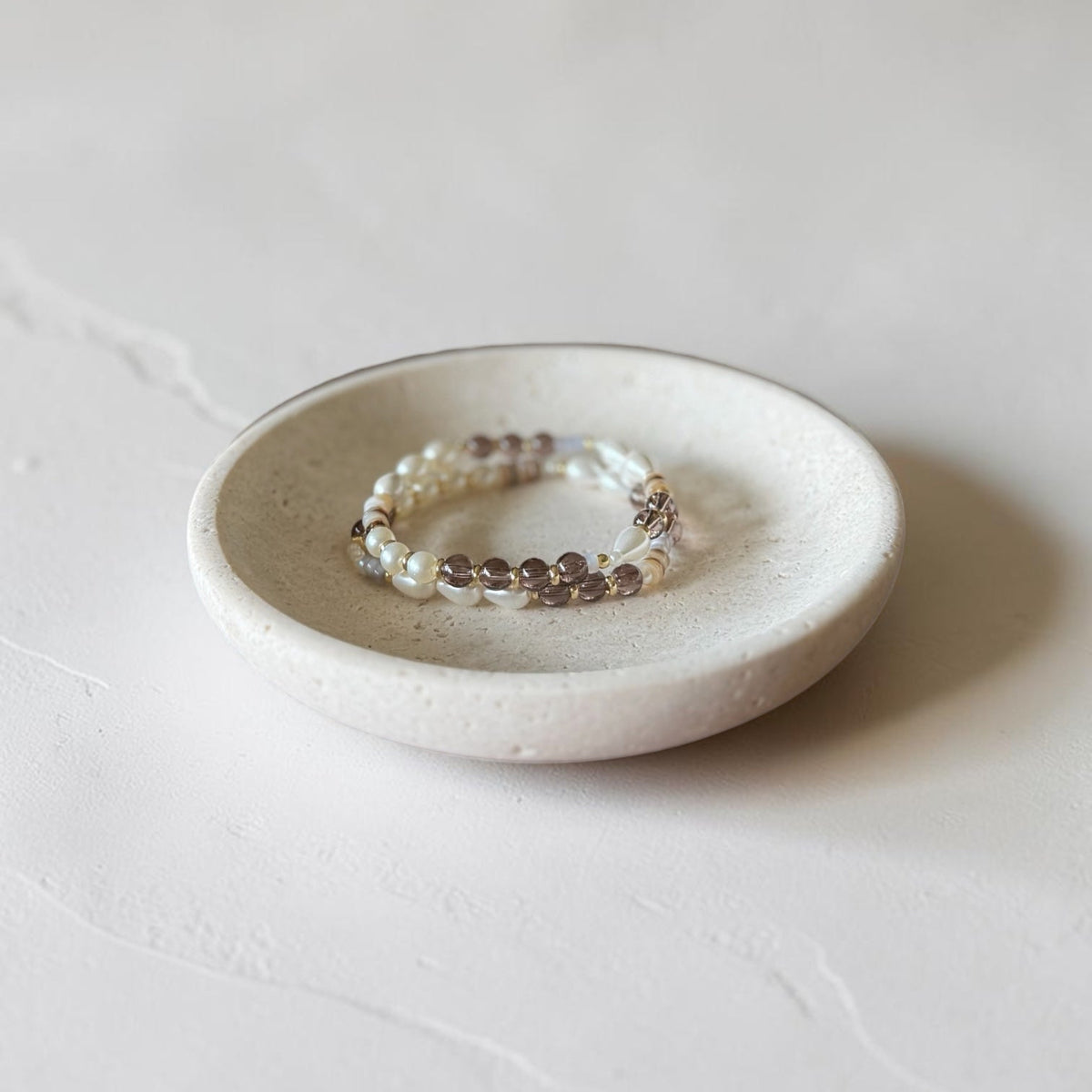 A beaded stretch bracelet with white pearls and brown gemstone beads resting on a small, round, textured ceramic dish against a light gray background.