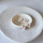 A pair of delicate gold hoop earrings with small clusters of white faux pearls, displayed on a small round ceramic dish resting on a larger speckled ceramic plate.