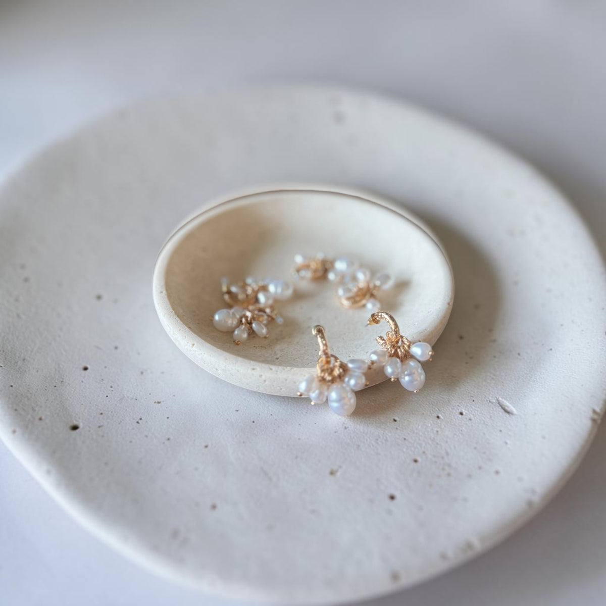 A pair of delicate gold hoop earrings with small clusters of white faux pearls, displayed on a small round ceramic dish resting on a larger speckled ceramic plate.