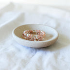 A small round ceramic dish holding a stack of beaded stretch bracelets in soft pink and white tones, set on a white fabric surface.