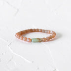 A beaded stretch bracelet featuring peachy-pink round beads and a central light green rectangular stone accent, displayed on a textured white surface.